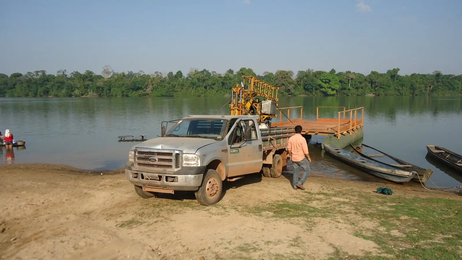 Caminhão da Geosul com equipamento de sondagem sendo embarcado em balsa às margens de um rio, cercado de mata