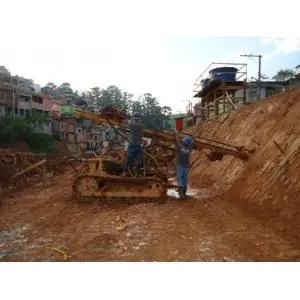 Perfuratriz sobre esteiras instalando grampos em talude de corte, em encosta urbana ocupada, com casas ao fundo